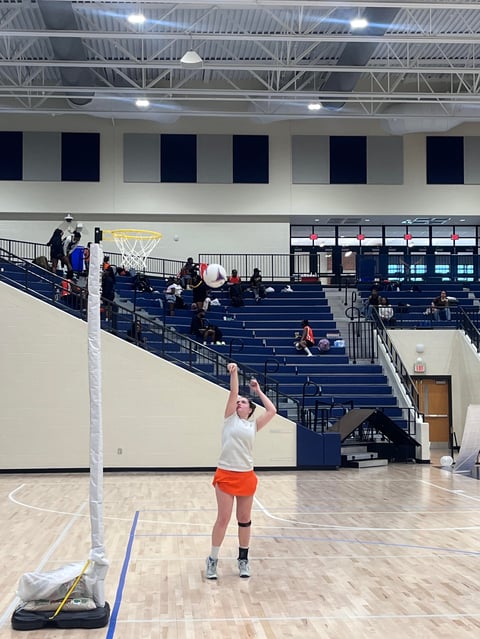 Volleyball player in white shirt and orange shorts serving a ball indoors on a wooden court with blue bleachers and basketball hoop visible