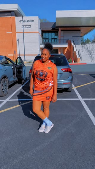 Young girl in orange outfit standing in parking lot in front of gymnasium building on sunny day