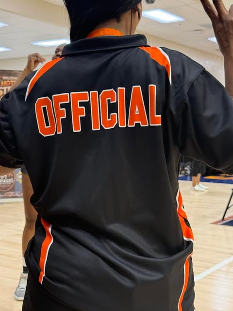 Person wearing a black and orange jacket with OFFICIAL text on the back, standing in an indoor gymnasium setting