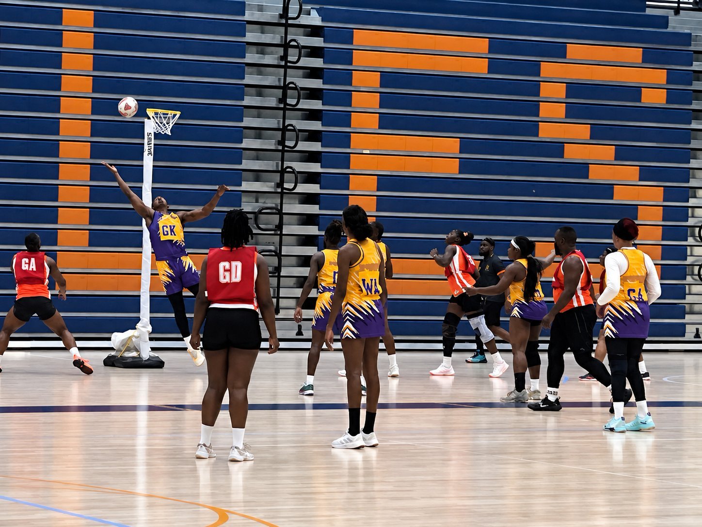 Netball players in yellow and red uniforms on indoor court with blue and orange striped wall during practice or match