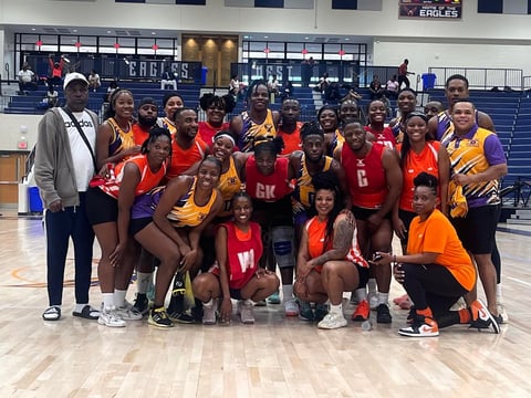 Group photo of a female volleyball team in red and gold uniforms posing together on an indoor court with bleachers in background