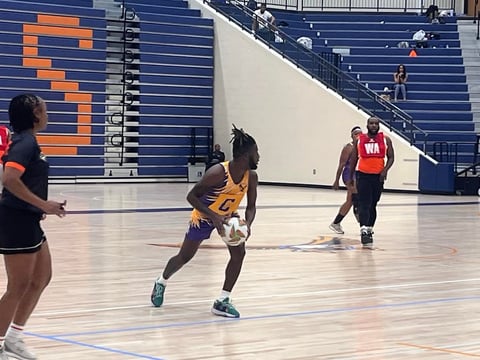 Netball player in purple and gold jersey passing the ball during a game in an indoor stadium with blue bleachers