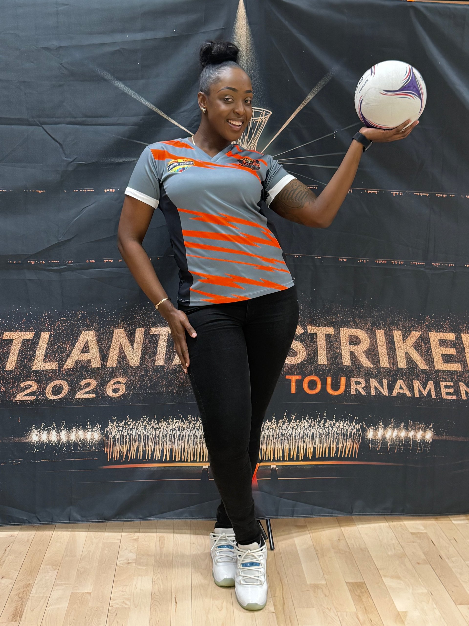 Young female soccer player in orange and gray jersey holding a soccer ball in front of Atlanta Strikers 2026 tournament banner